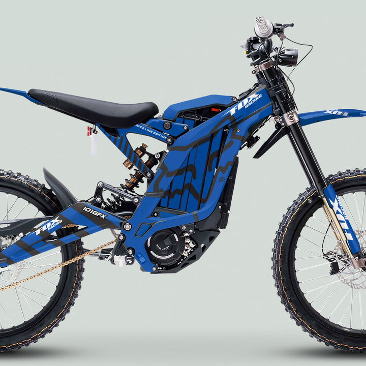 Blue electric dirt bike with Surron Light Bee X Graphics Kit FOX RACE LINE, featuring knobbly tires, electric motor, and striking blue-black design.